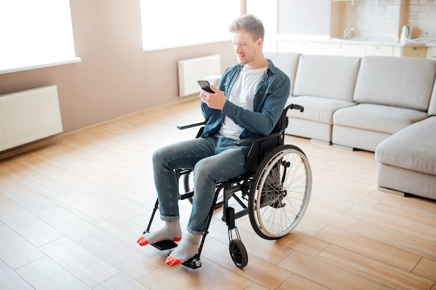 Netter junger Mann mit Inklusion und Behinderung. Im Rollstuhl sitzen. Telefon in Händen halten und anschauen. Tageslicht in einem großen leeren Raum.