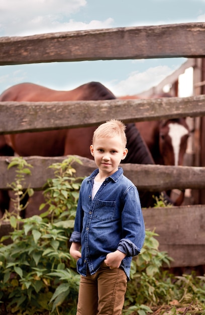 Netter Kindercowboy, der in der Nähe von Holzzaun mit Pferden steht