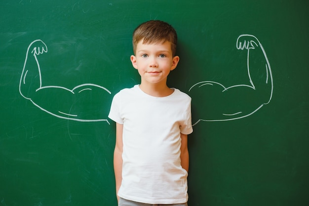 Netter Kinderjunge in Schuluniform und Brille. Gehe zum ersten Mal zur Schule. Kind mit Schultasche und Büchern. Kind im Klassenzimmer in der Nähe der Tafel mit Muskeln darauf. Zurück zur Schule