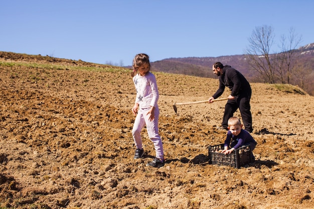 Netter kleiner blonder Junge, der seiner Familie hilft, Kartoffeln im zeitigen Frühjahr zu pflanzen Landwirtschaftliche Arbeiten auf dem Bauernhof hoch in den Bergen Schöne schneebedeckte Bergspitze auf einem Hintergrund