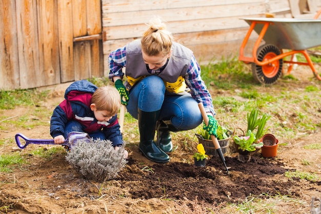 Netter kleiner Gärtner mit blonden Haaren, der seiner Mutter hilft, Blumen in ihrem Garten zu pflanzen. Junge mit Spielzeugschaufel, der im Boden gräbt und vorgibt, Gärtner zu sein, Helfer der Mutter