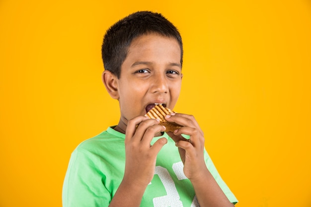 Netter kleiner indischer oder asiatischer Junge, der leckeren Burger, Sandwich oder Pizza in einem Teller oder einer Schachtel isst. Stehend lokalisiert über blauem oder gelbem Hintergrund.