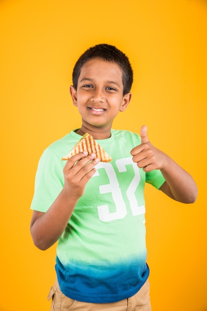 Netter kleiner indischer oder asiatischer Junge, der leckeren Burger, Sandwich oder Pizza in einem Teller oder einer Schachtel isst. Stehend lokalisiert über blauem oder gelbem Hintergrund.