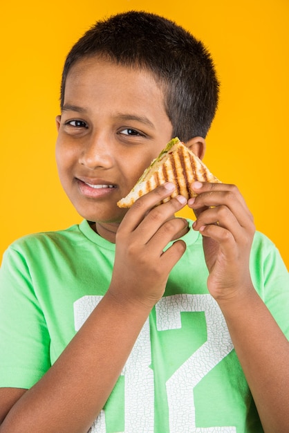 Netter kleiner indischer oder asiatischer Junge, der leckeren Burger, Sandwich oder Pizza in einem Teller oder einer Schachtel isst. Stehend lokalisiert über blauem oder gelbem Hintergrund.