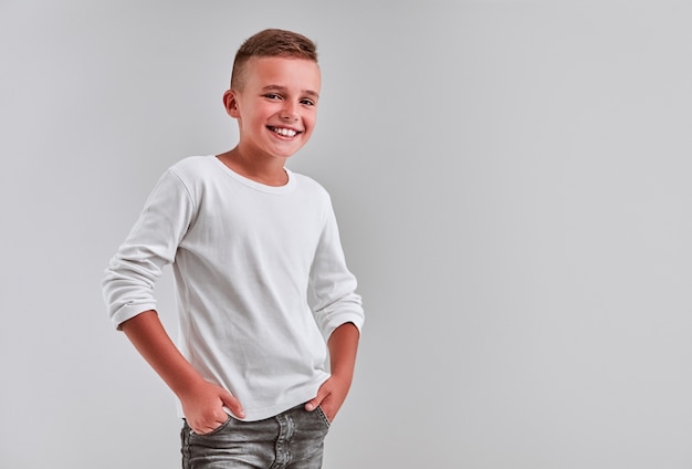 Foto netter kleiner junge auf grauem hintergrund hält seine hände in seinen jeanstaschen und lächelt