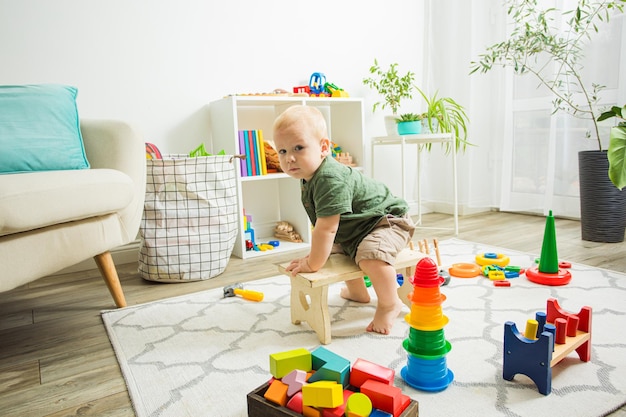 Netter kleiner Junge, der auf einer kleinen Holzbank sitzt und wie ein Pferd reitet Kleiner Junge, der vorgibt, ein Cowboy zu sein Kinderzimmer mit vielen bunten Spielsachen