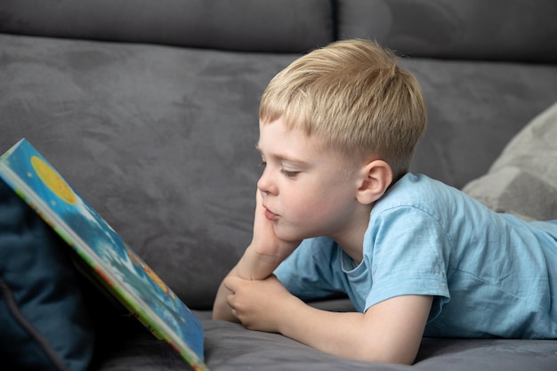 Netter kleiner Junge, der ein Buch liest, das auf der Couch liegt Der Junge interessiert sich für das Konzept der Bildung