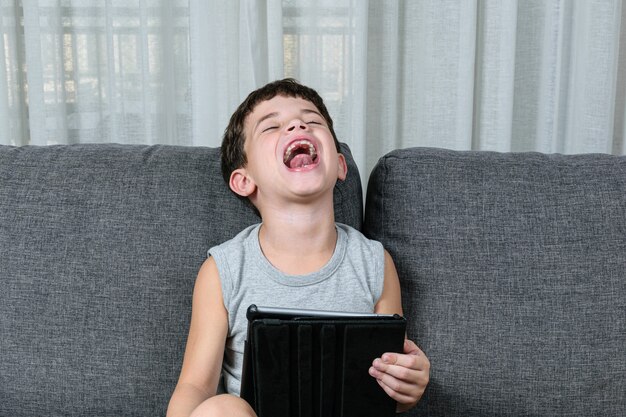 Netter kleiner Junge, der eine Tablette auf dem Sofa benutzt