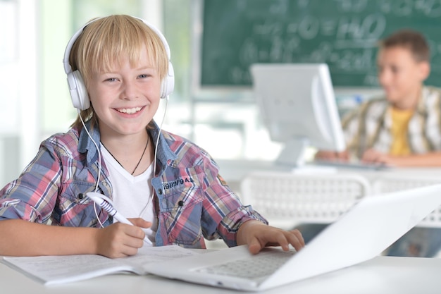 Netter kleiner Junge, der mit Laptop am Tisch sitzt, während er Hausaufgaben macht und Daumen hoch zeigt