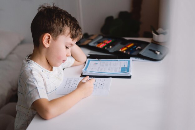 Netter kleiner Junge, der zu Hause lernt und Schulhausaufgaben macht und denkt, dass das Kind am Tisch sitzt