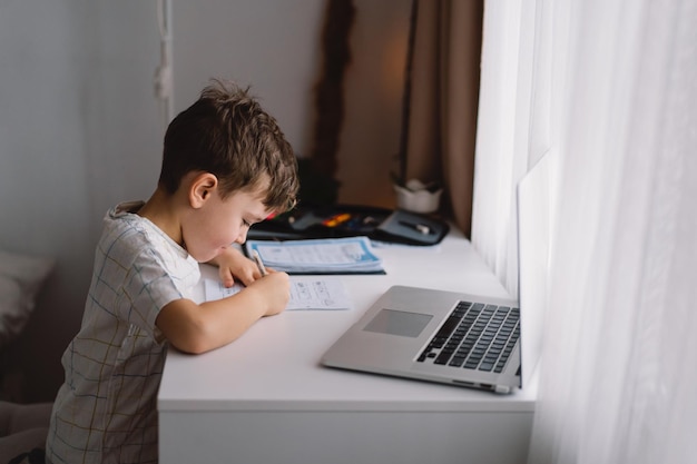 Netter kleiner Junge, der zu Hause mit Laptop lernt und Schulhausaufgaben macht