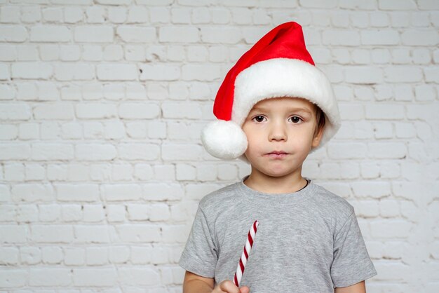 Netter kleiner Junge in der Weihnachtsmütze, die Weihnachtszuckerrohrbonbons auf weißem Backsteinwandhintergrund isst