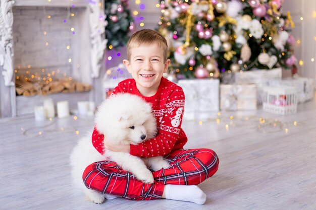 Netter kleiner Junge mit einem weißen Samojeden-Hündchen nahe dem Weihnachtsbaum, Feiertagskonzept, neues Jahr