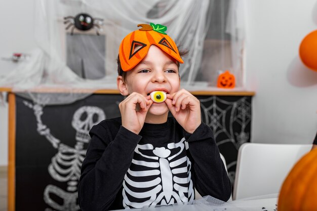 Netter kleiner Junge mit gruseligem Kostüm, das seine Halloween-Süßigkeiten genießt. Jack O 'Lantern Halloween Kürbis auf dem Tisch und andere gruselige Dekorationen