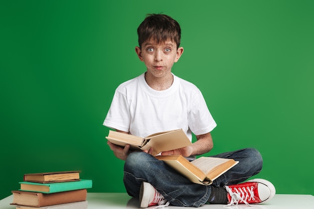 Netter kleiner Junge mit Sommersprossen, der mit Bücherstapeln über grüner Wand sitzt