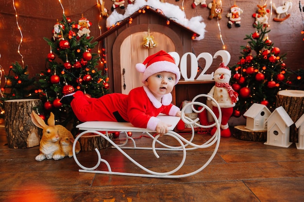 Netter kleiner Junge verkleidet als Weihnachtsmann auf einem alten Vintage-Schlitten mit Geschenken in der Nähe des Weihnachtsbaums, fröhliches Urlaubskonzept. 2022