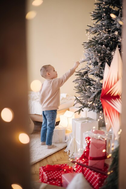 Netter kleiner Junge zu Hause während der Winterferien Junge geschmückter Weihnachtsbaum und viele Geschenke