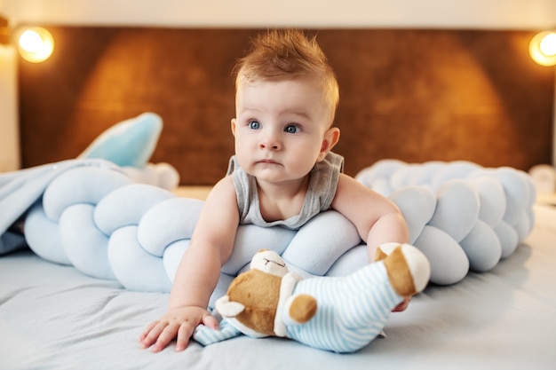 Netter kleiner kaukasischer Junge, der auf Bauch auf med liegt, mit Teddybär spielt und neugierig schaut.