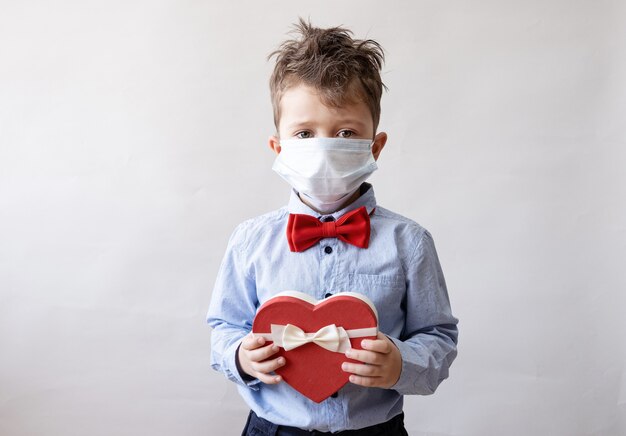 Netter kleiner kaukasischer Junge in Fliege mit weißem Band der roten Herzgeschenkbox in der Gesichtsmaske. Valentinstag. Covid.