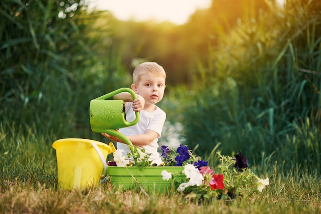 Netter kleiner Kinderjunge, der Blumensämlinge in einem Topf im Garten auf Sonnenuntergang wässert. Lustiger kleiner Gärtner. Frühlingskonzept, Natur und Pflege.