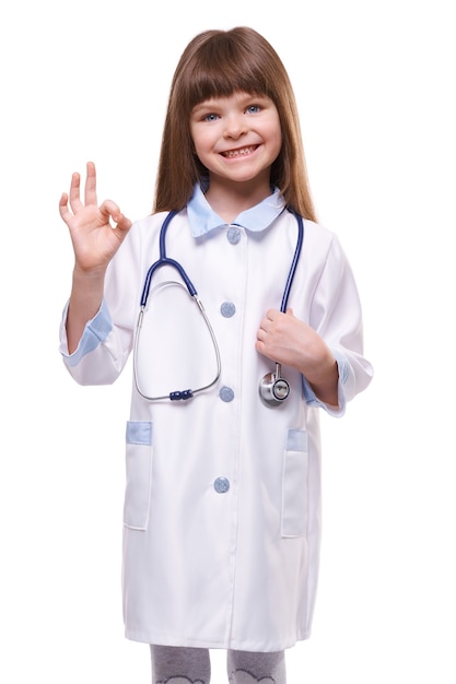 Netter kleiner Mädchenarzt, der weißen Kittel trägt, hält Stethoskop und zeigt OK-Geste auf weißem lokalisiertem Hintergrund
