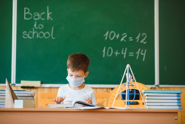 Netter kleiner Schuljunge, der im Klassenzimmer mit einer Maske sitzt