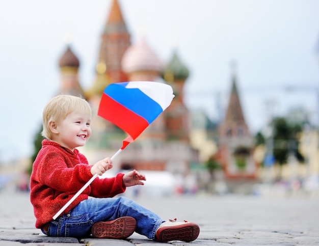 Netter Kleinkindjunge, der mit russischer Flagge mit Abstammung des Roten Platzes und Vasilevsky sitzt und spielt