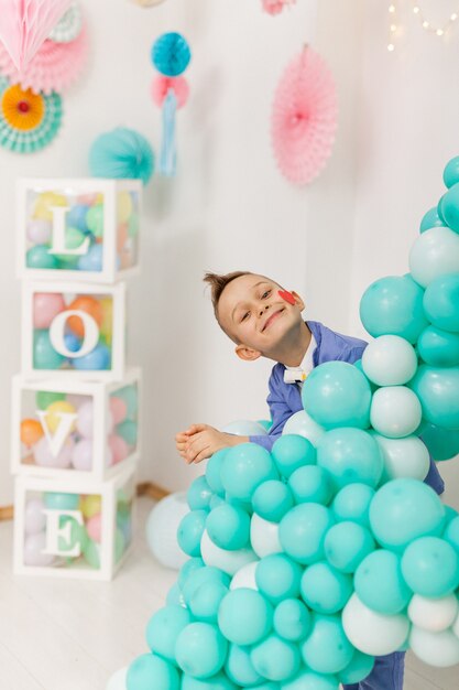 Netter lachender Junge mit einem roten Herzen auf seiner Wange, das hinter bunten Partyballons hervorschaut