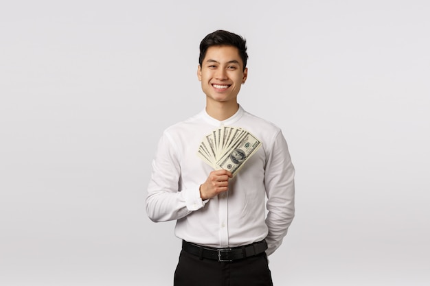 Netter lächelnder asiatischer junger Unternehmer mit dem weißen Hemd, das Banknoten hält