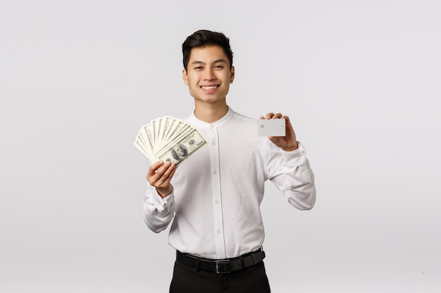 Netter lächelnder asiatischer junger Unternehmer mit dem weißen Hemd, das Kreditkarte und Banknoten hält