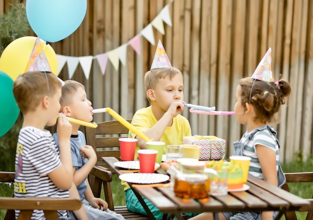 Netter lustiger neunjähriger Junge, der seinen Geburtstag mit Familie oder Freunden mit hausgemachtem gebackenem Kuchen in einem Hinterhof feiert Geburtstagsfeier Kinder, die Partyhüte tragen und Pfeifen blasen