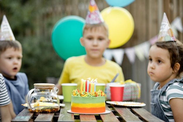 Netter lustiger neunjähriger Junge, der seinen Geburtstag mit Familie oder Freunden mit hausgemachtem gebackenem Kuchen in einem Hinterhof feiert Geburtstagsfeier Kinder, die Partyhüte tragen