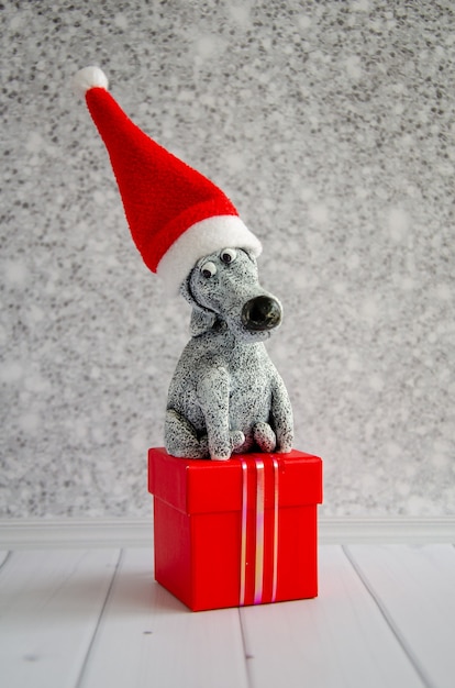 Netter lustiger Spielzeughund mit Weihnachtsmann-Hut sitzt auf roter Geschenkbox vor dem Hintergrund der glänzenden grauen Wand.