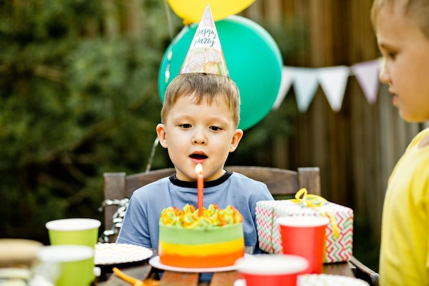 Netter lustiger vierjähriger Junge, der seinen Geburtstag mit Familie oder Freunden feiert, die Kerzen auf hausgemachtem gebackenem Kuchen in einer Hinterhof-Geburtstagsfeier für Kinder blasen