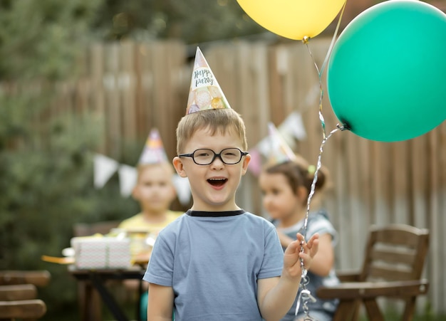 Netter lustiger vierjähriger Junge, der seinen Geburtstag mit Familie oder Freunden in einem Hinterhof feiert Geburtstagsfeier Kind mit Partyhut und Ballon in der Hand