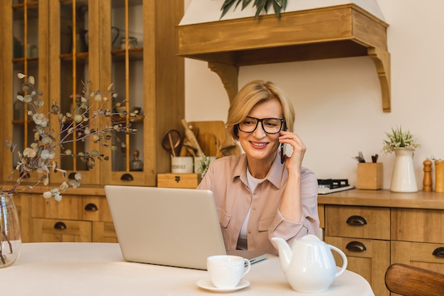 Netter Morgen. Fröhlich lächelnde ältere Frau, die in der Küche steht und ihren Laptop benutzt, während sie sich nach dem Frühstück ausruht, Freiberuflerin, die zu Hause arbeitet. Handy benutzen.