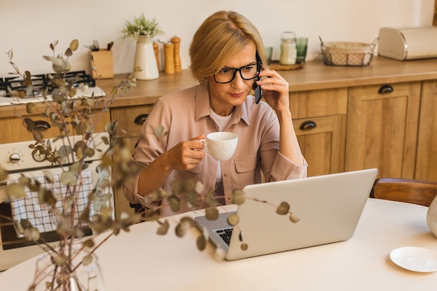 Netter Morgen. Fröhlich lächelnde ältere Frau, die in der Küche steht und ihren Laptop benutzt, während sie sich nach dem Frühstück ausruht, Freiberuflerin, die zu Hause arbeitet. Handy benutzen.