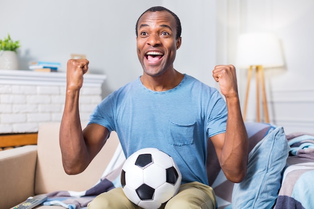 Netter positiver glücklicher Mann, der einen Ball hält und sein Glück ausdrückt, während er über seine Fußballmannschaft glücklich ist