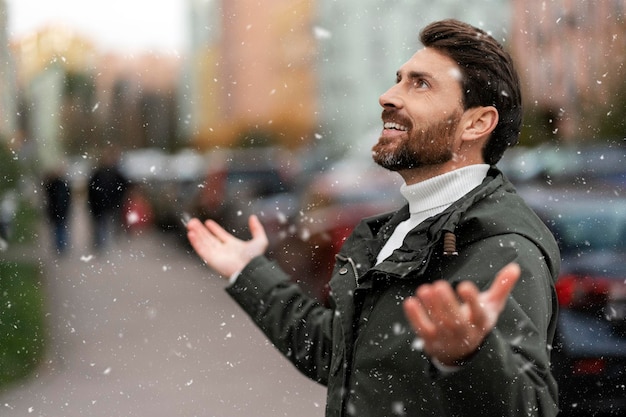 Netter positiver Mann im Freien, der Schneefall beobachtet und nach oben schaut Ein Mann vor dem Hintergrund der Stadt