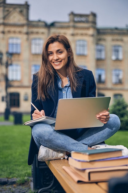 Netter Student, der mit gekreuzten Beinen auf der Bank sitzt und lächelt, während er am Laptop arbeitet