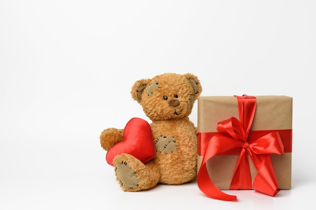 Netter Teddybär, der ein rotes Herz hält, neben einer Schachtel mit einem Geschenk, das mit einem roten Seidenband, weißer Hintergrund gebunden wird