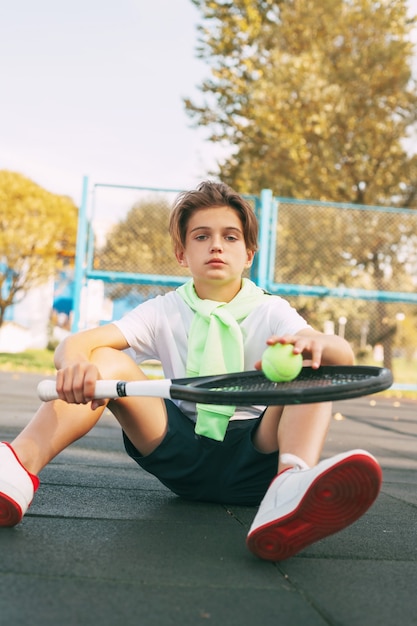 Foto netter teenager, der auf dem tennisplatz sitzt und sich nach einem training ausruht. sport, sportler, lebensstil