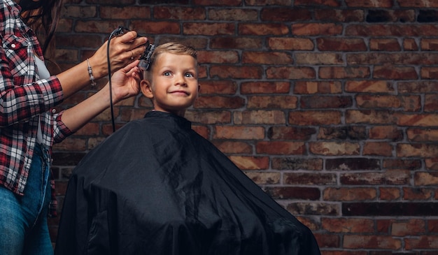 Netter Vorschuljunge, der Haarschnitt bekommt. Kinderfriseur mit Trimmer schneidet kleinen Jungen im Raum mit Loft-Interieur.