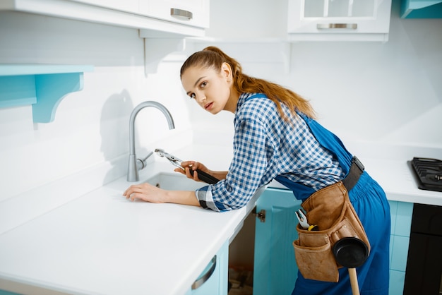 Netter weiblicher Klempner im einheitlichen Befestigungshahn in der Küche. Heimwerkerin mit Werkzeugtaschen-Reparaturspüle, Sanitäranlagen-Service zu Hause