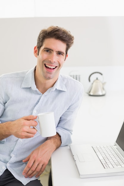 Netter zufälliger Mann mit Kaffeetasse mit Laptop zu Hause