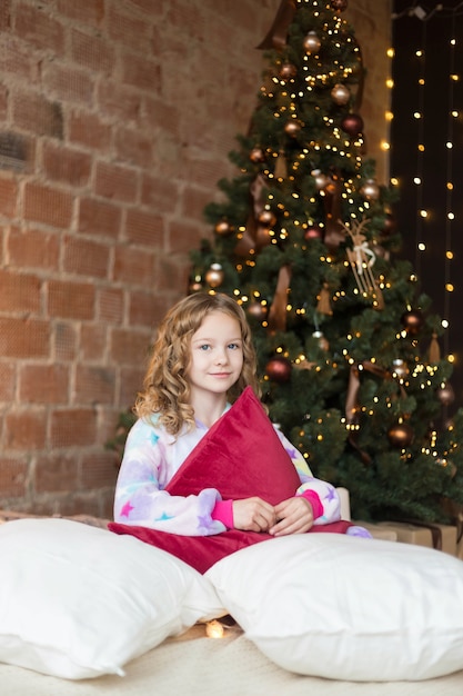 Nettes 10 Jahre altes Mädchen sitzt auf dem Bett und umarmt Kissen mit Weihnachtsbaumverhalten