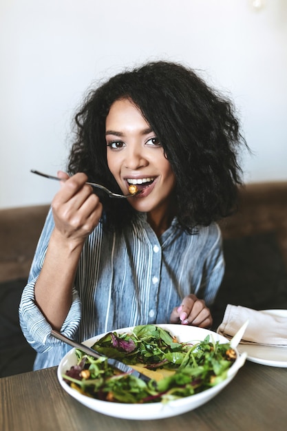 Nettes afroamerikanisches Mädchen, das Salat im Restaurant isst. Porträt der lächelnden Dame mit dunklem lockigem Haar und Salat auf Tisch am Café