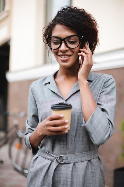 Nettes afroamerikanisches Mädchen in den Gläsern, die auf Straße stehen und auf ihrem Handy mit Kaffee sprechen