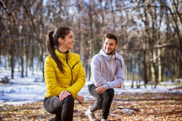 Nettes aktives Fitnessmädchen in der Sportbekleidung, die Beinstrecken mit ihrem persönlichen Trainer im Wald am sonnigen Wintermorgen tut.