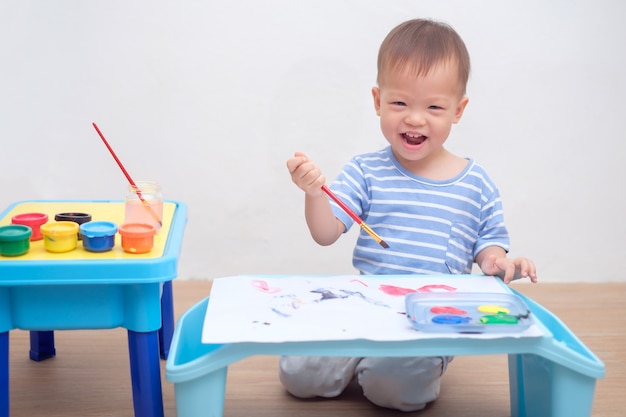 Nettes asiatisches 1-jähriges Kleinkindjungenkind, das mit Pinsel u. Aquarellen zu Hause malt, kreative Kunstaktivitäten für körperliche Entwicklung, großes und kleines Muskelentwicklungskonzept der Kinder